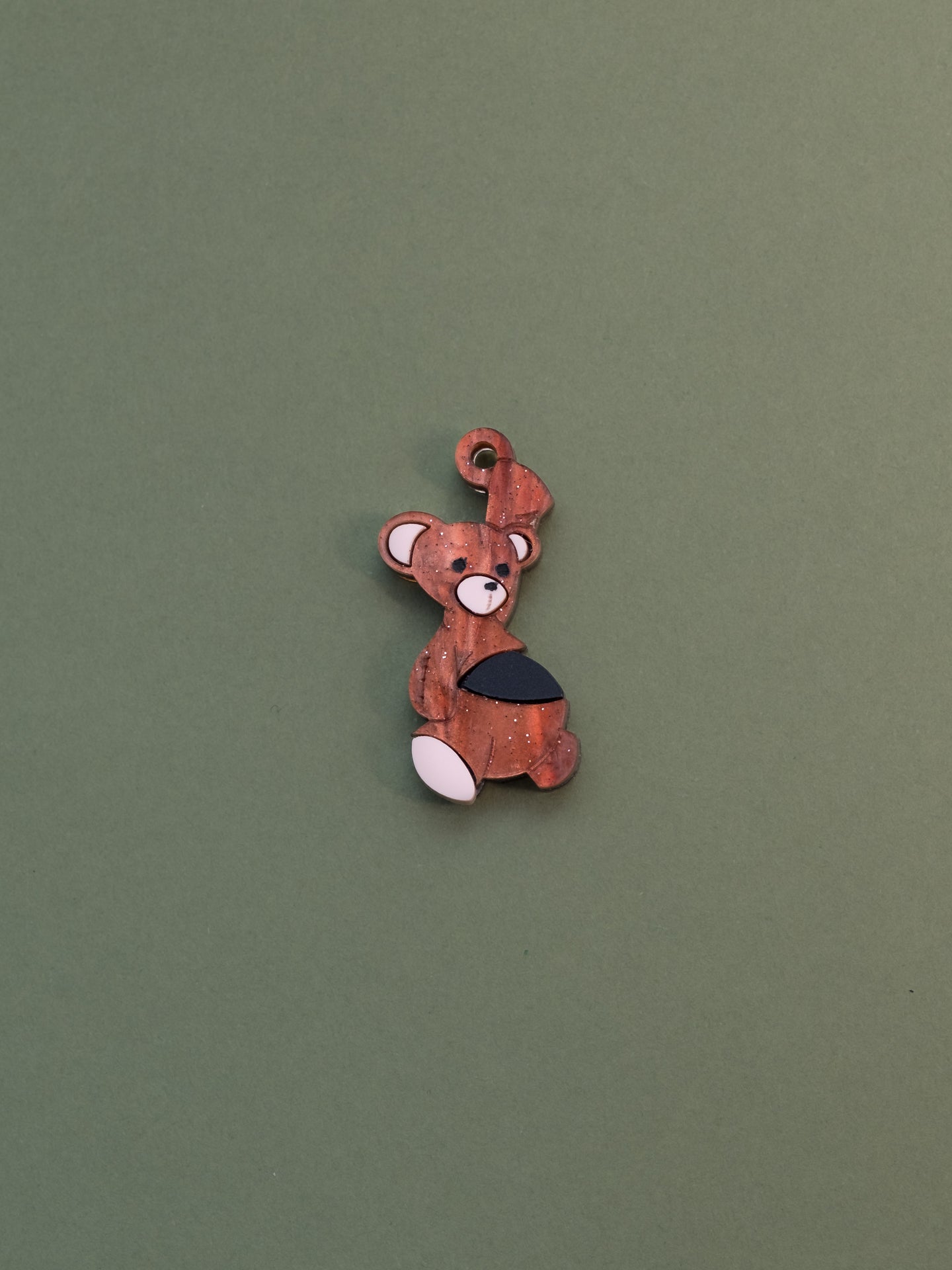 Charm de metacrilato inspirado en el osito de peluche de Michael de peter pan, con una bala de cañon en la barriga, en color marrón aguas con purpurina y la bala en negro
