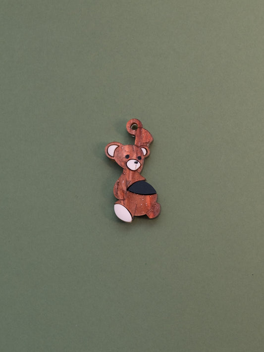 Charm de metacrilato inspirado en el osito de peluche de Michael de peter pan, con una bala de cañon en la barriga, en color marrón aguas con purpurina y la bala en negro